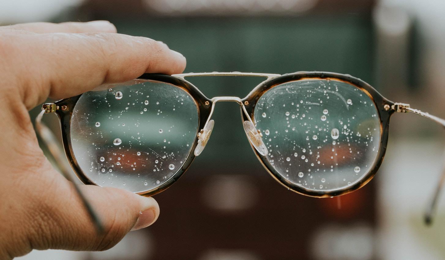 image of a hand holding smeared glasses covered in rain drops