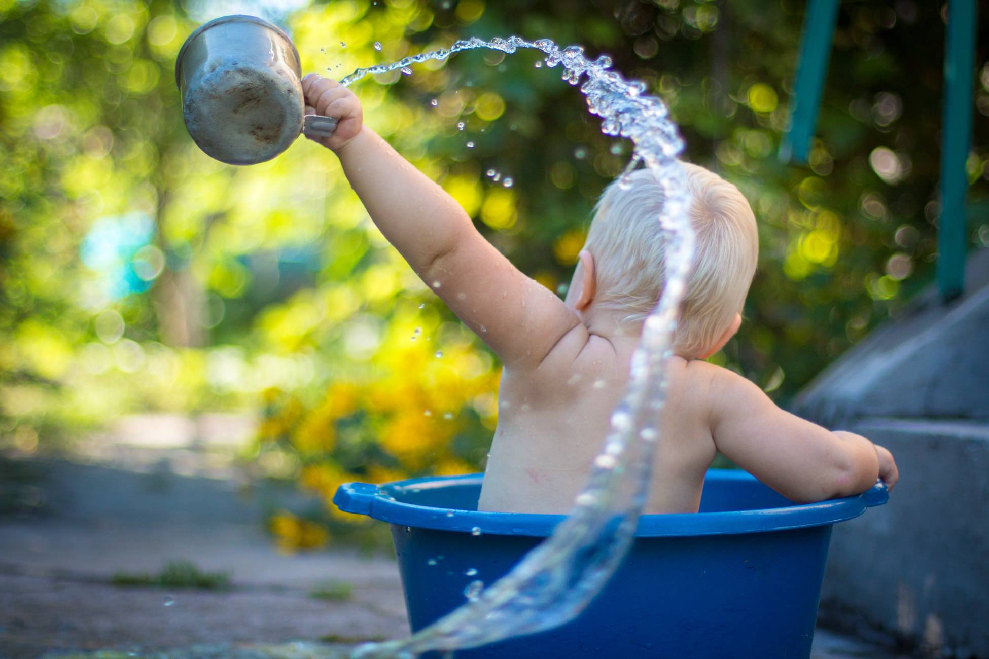 lifestyle photography shot of a little boy in a bucket of water throwing a cup of water in the air